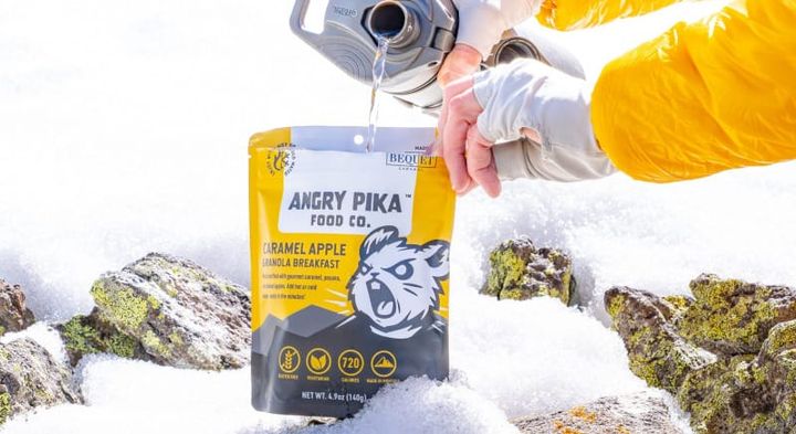 Cold water being poured into an Angry Pika Caramel Apple Granola Breakfast pouch in a snowy alpine setting.