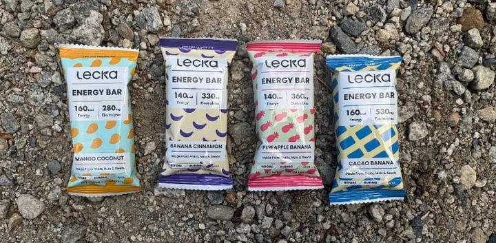 Four Lecka Energy Bars—Mango Coconut, Banana Cinnamon, Pineapple Banana, and Cacao Banana—arranged on rocky ground.