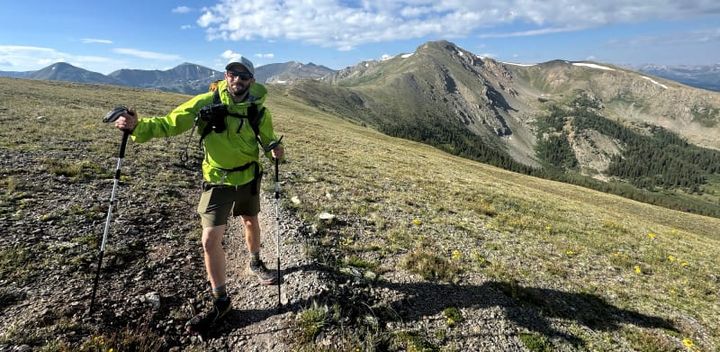  Hiker wearing HIMALI Monsoon Hardshell Jacket with trekking poles walking through an alpine meadow with a dramatic ridgeline in the distance.