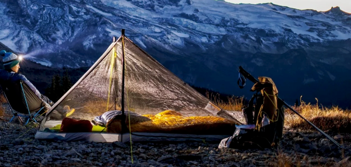 Backpacker relaxing next to a lit Six Moon Designs ultralight tent with sleeping bag inside, set up in a rugged alpine landscape at dusk with snowy mountains in the background.