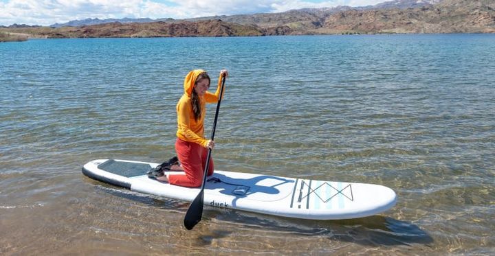  Woman kneeling on a paddleboard in shallow clear water, wearing a bright orange Wuru Wool Lightweight Hoodie with red pants, surrounded by desert mountains and a blue sky.