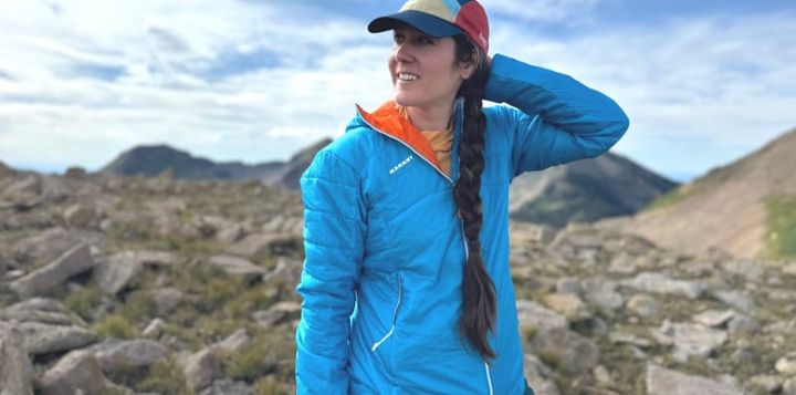 Hiker wearing the Mammut Eigerjoch Light IN Hooded Jacket in bright blue while exploring a rocky alpine ridge under a partly cloudy sky.
