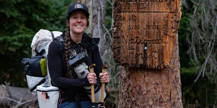 Backpacker with trekking poles standing beside a weathered wilderness trail sign in the San Juan Mountains.