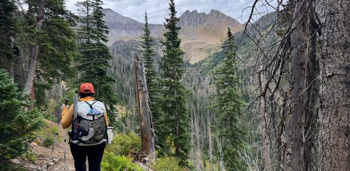 Hiker wearing the Hyperlite Windrider 70 Backpack while trekking through alpine forest toward rocky peaks.