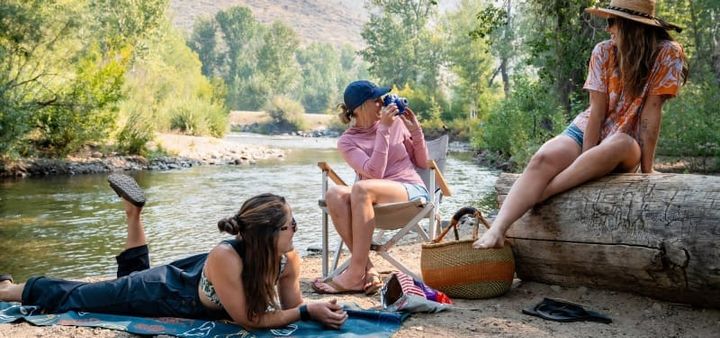  Three women enjoy a sunny afternoon by a river, lounging on a blanket, sitting in a camp chair, and perching on a log, all wearing colorful and comfortable Wild Rye clothing.