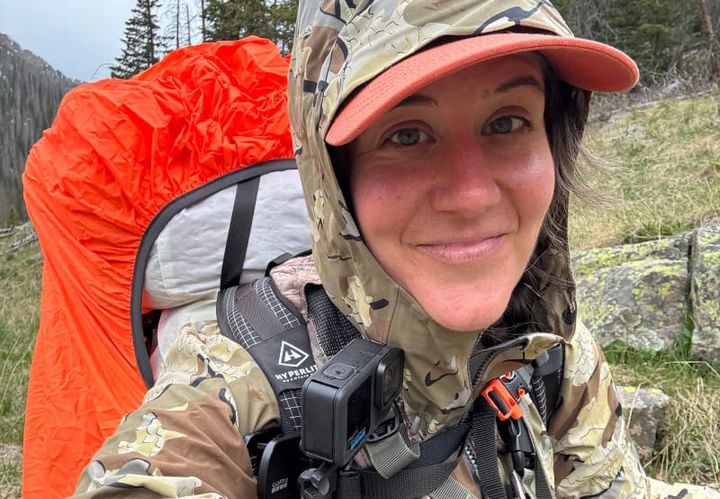 Backpacker wearing KUIU camo rain jacket with hood up, hiking through wet mountain terrain with a bright orange pack cover, showcasing durable rain protection.