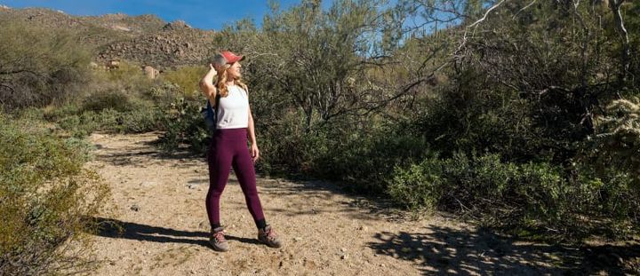 Hiker in a red cap standing on a desert trail lined with brush and mountains in the distance beneath a bright blue sky.