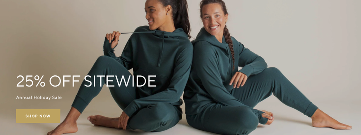 Two women wearing matching teal BRANWYN Merino loungewear sets sit on the floor smiling and relaxing during the brand’s annual Holiday Sale offering 25% off sitewide.