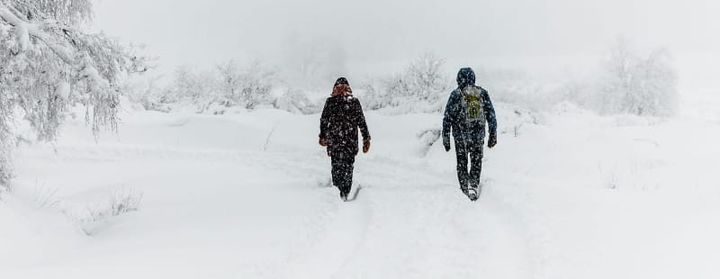 Two hikers walking through deep fresh snow on a foggy winter day, surrounded by snow-covered trees and a soft white landscape.