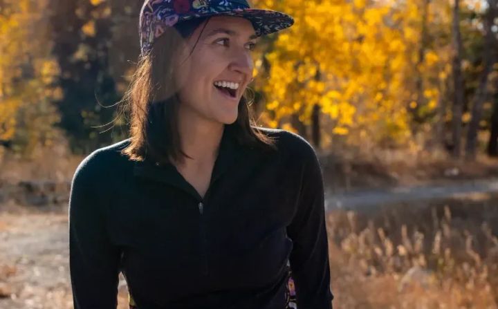 Woman wearing the Wild Rye Bassett Half Zip in black, smiling outdoors with golden fall trees behind her.