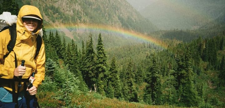 Hiker wearing a yellow rain jacket with trekking poles on a mountain trail as a rainbow arcs over forested peaks