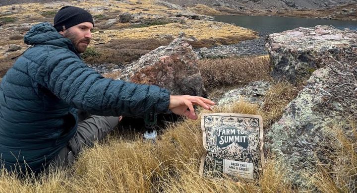  backpacker reaching for Farm to Summit dehydrated backpacking meal displayed on a rock surface, featuring compostable packaging and locally sourced ingredients.