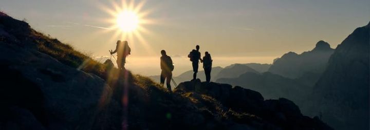 Group of hikers standing on a mountain ridge at sunrise with bright sunlight over an alpine landscape.
