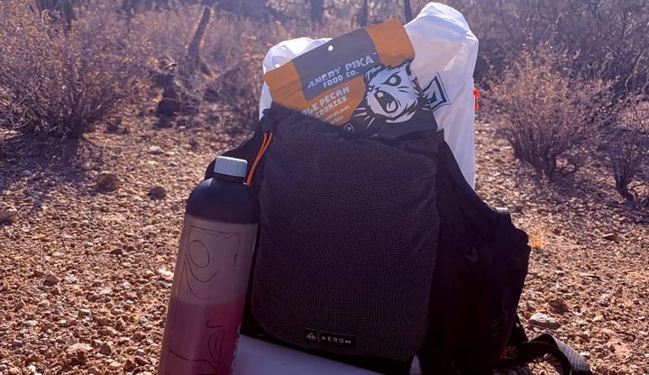 Hyperlite Mountain Gear Aero 28 ultralight fastpacking backpack with water bottles and trail snacks on a rocky Sonoran Desert hiking trail surrounded by saguaros.