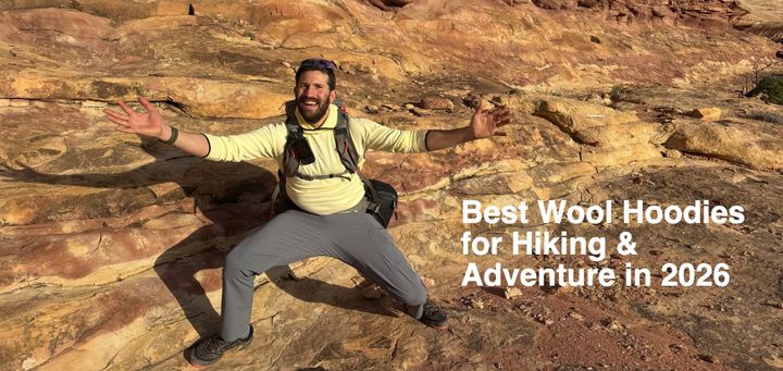 Hiker wearing a pale yellow hoodie and backpack, sitting on a sandstone ledge with arms outstretched beneath a towering red rock overhang.