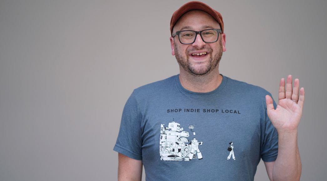 I am smiling and waving to the camera while wearing a Shop Indie Shop Local t-shirt designed by Adrian Tomine.