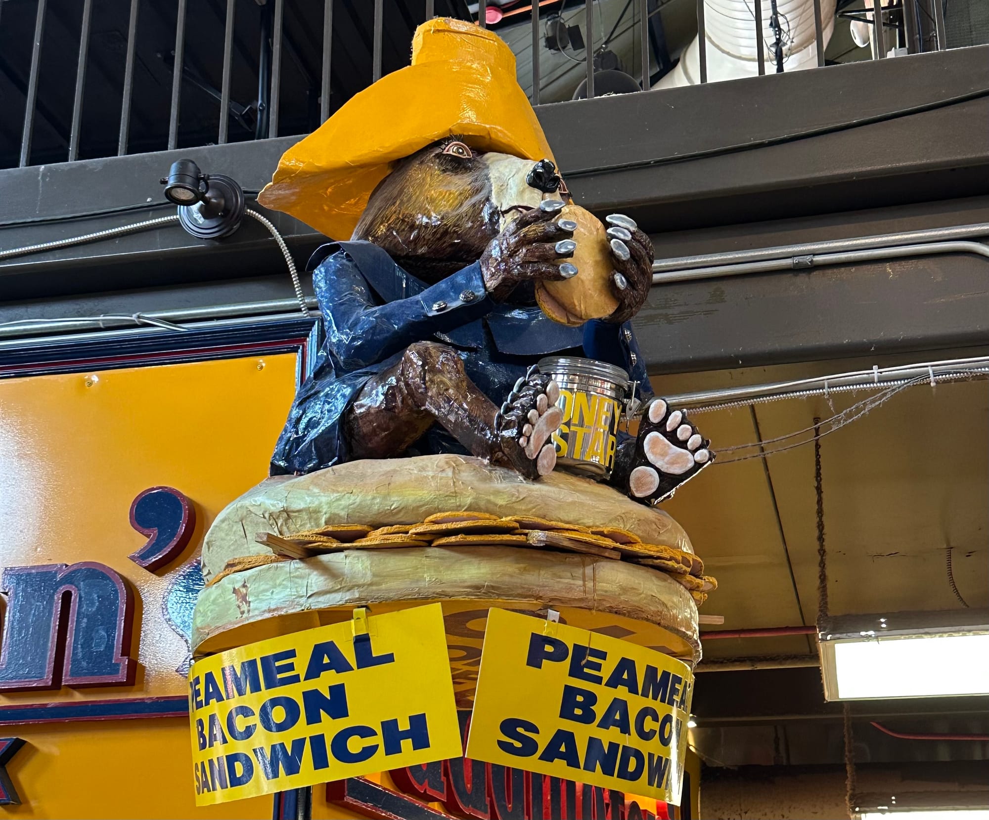 A wooden Paddington sculpture up in the air with signs advertising a "peameal bacon sandwich."