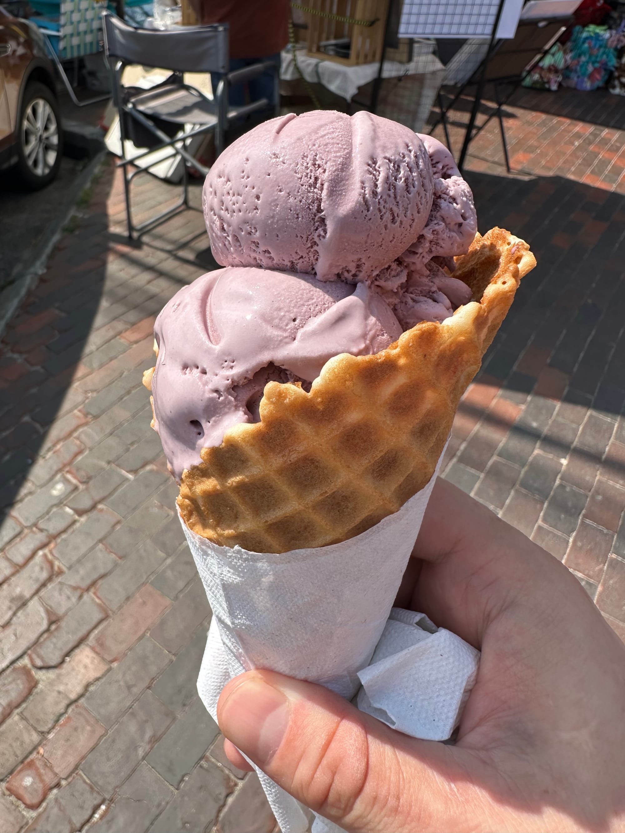 My hand holding a "small" waffle cone of black raspberry ice cream over a brick walkway. The cone is wrapped sensibly in a napkin.