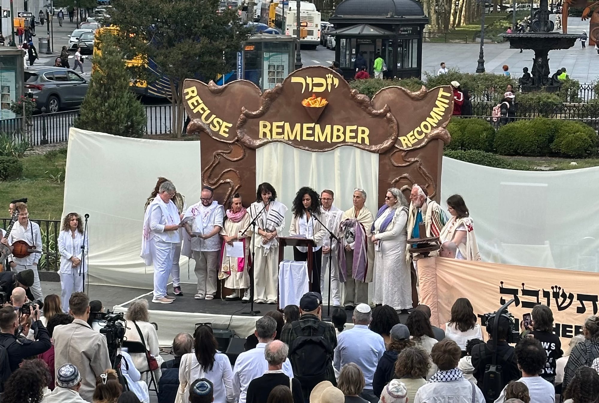 Hala Alyan speaking at a Yizkor service in Brooklyn surrounded by rabbis with some onlookers.