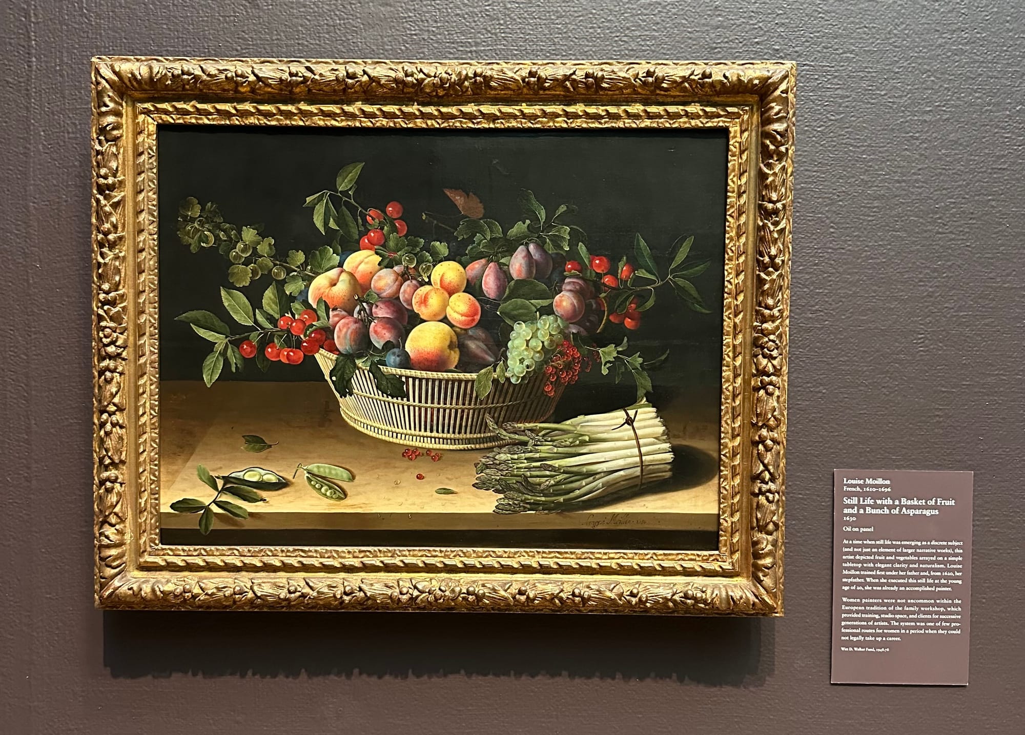 Still life full of fruits and veggies in an ornate gold-painted frame with the card identifying the French artist Louise Moillon.