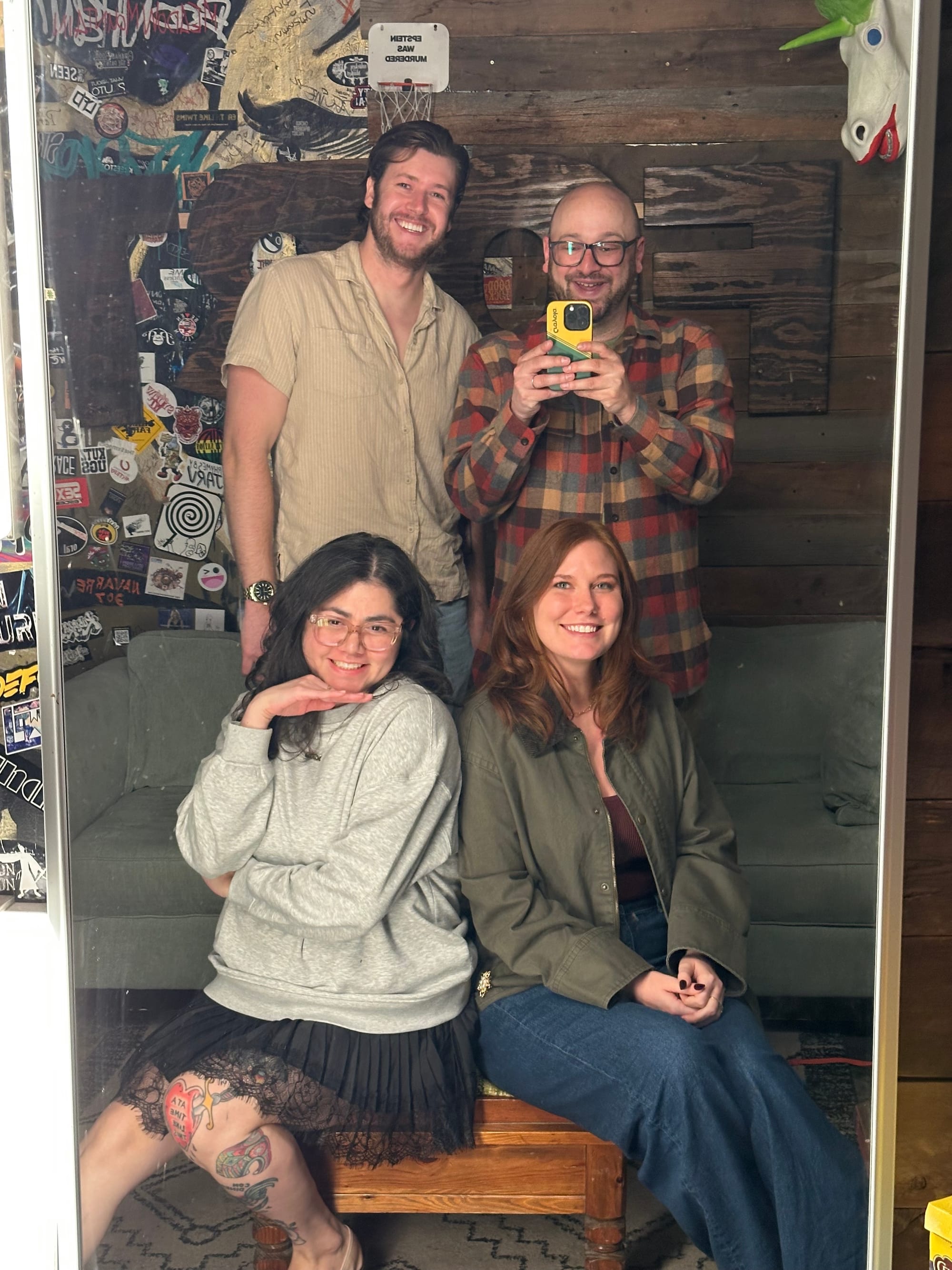 A mirror photo of me, Owen, Miriam, and Bailey at the Comedy Fort. Bailey and Miriam are sitting on an ottoman while Owen and I stand behind them.