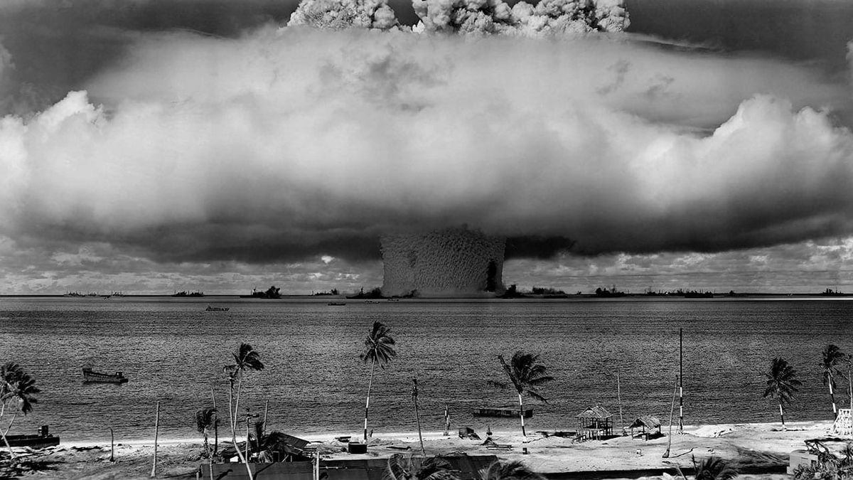 Nuclear weapons test in the Pacific. Giant mushroom cloud over the ocean with palm trees in the background.