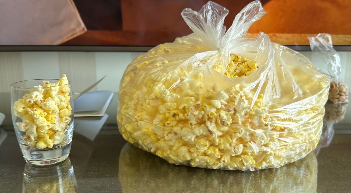 A giant plastic bag of popcorn torn open on top sitting beside a little water glass that's full of popcorn.