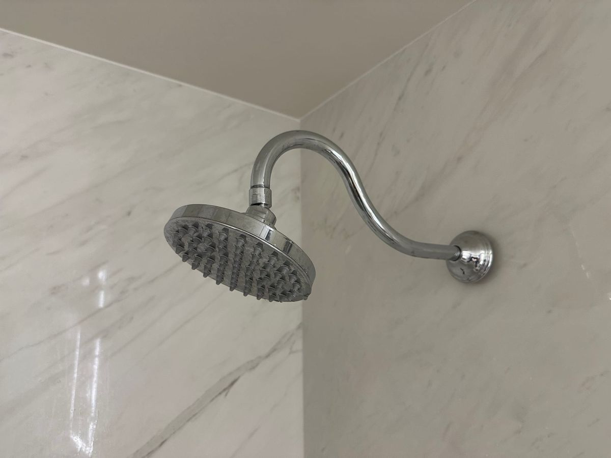 Disc-shaped shower head with lots of little spouts on a curvy pipe leading to a white marble looking wall.