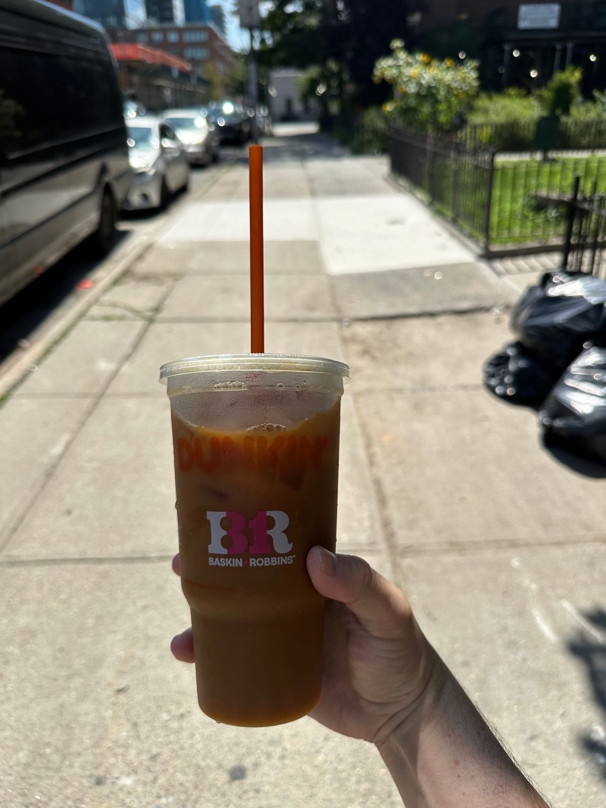 poorly lit dunkin iced coffee in the foreground with sidewalk and full black trash bags in the background