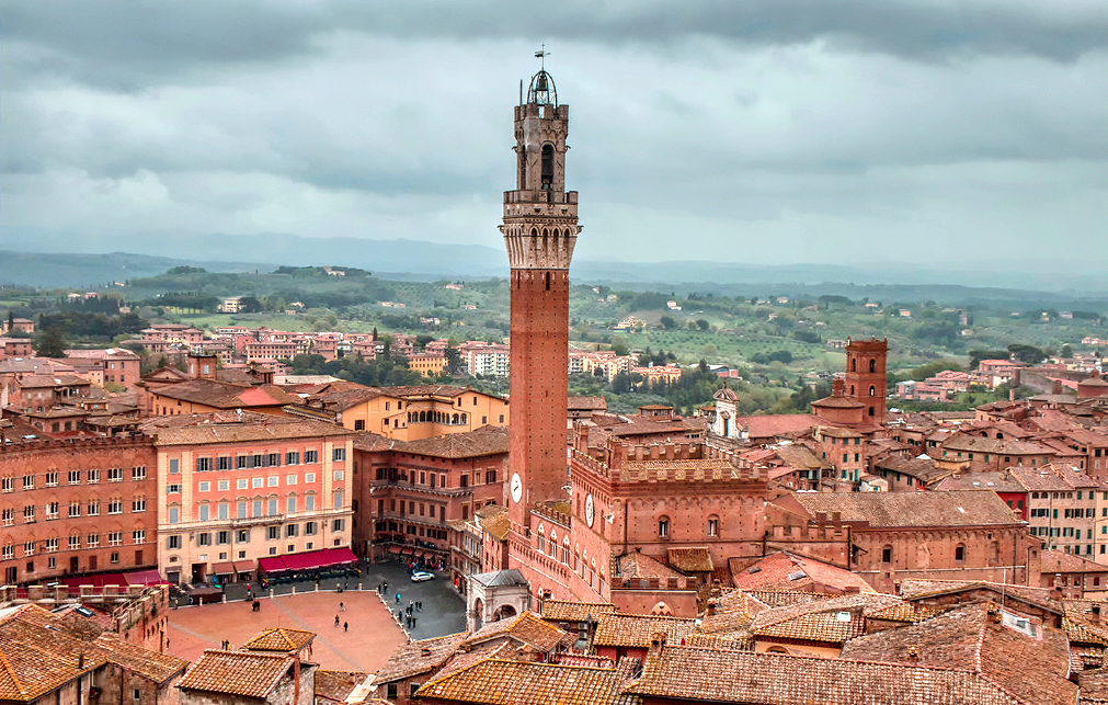 A wide panoramic view over the terracotta rooftops of Siena, capturing the sweeping curve of Piazza del Campo and the tall brick profile of the Torre del Mangia rising into the sky.