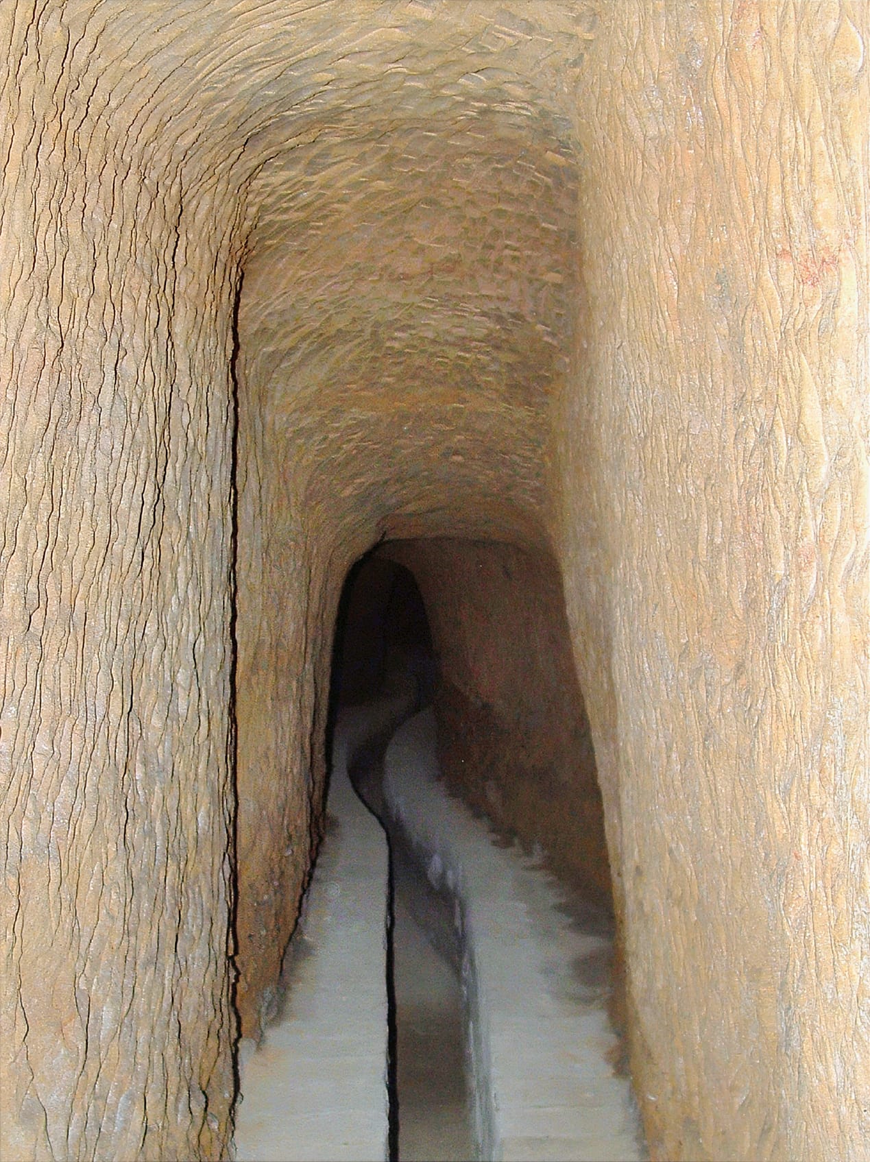 The narrow interior of an underground bottino aqueduct in Siena, carved through yellow tufa rock with a brick-vaulted ceiling.