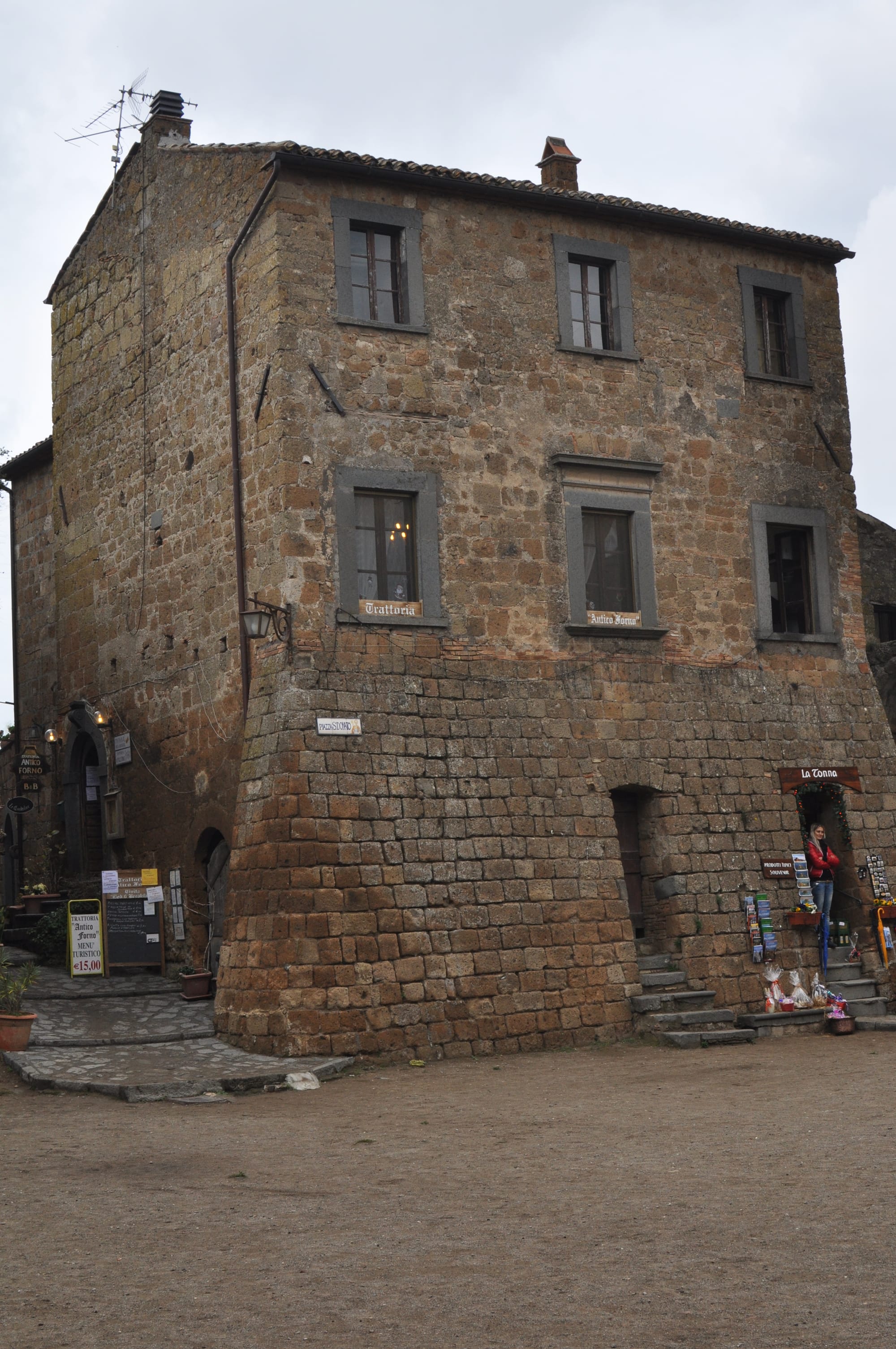 A prominent three-story medieval building constructed from rough tufa blocks, with signs for a local trattoria and stone steps leading to its entrances.