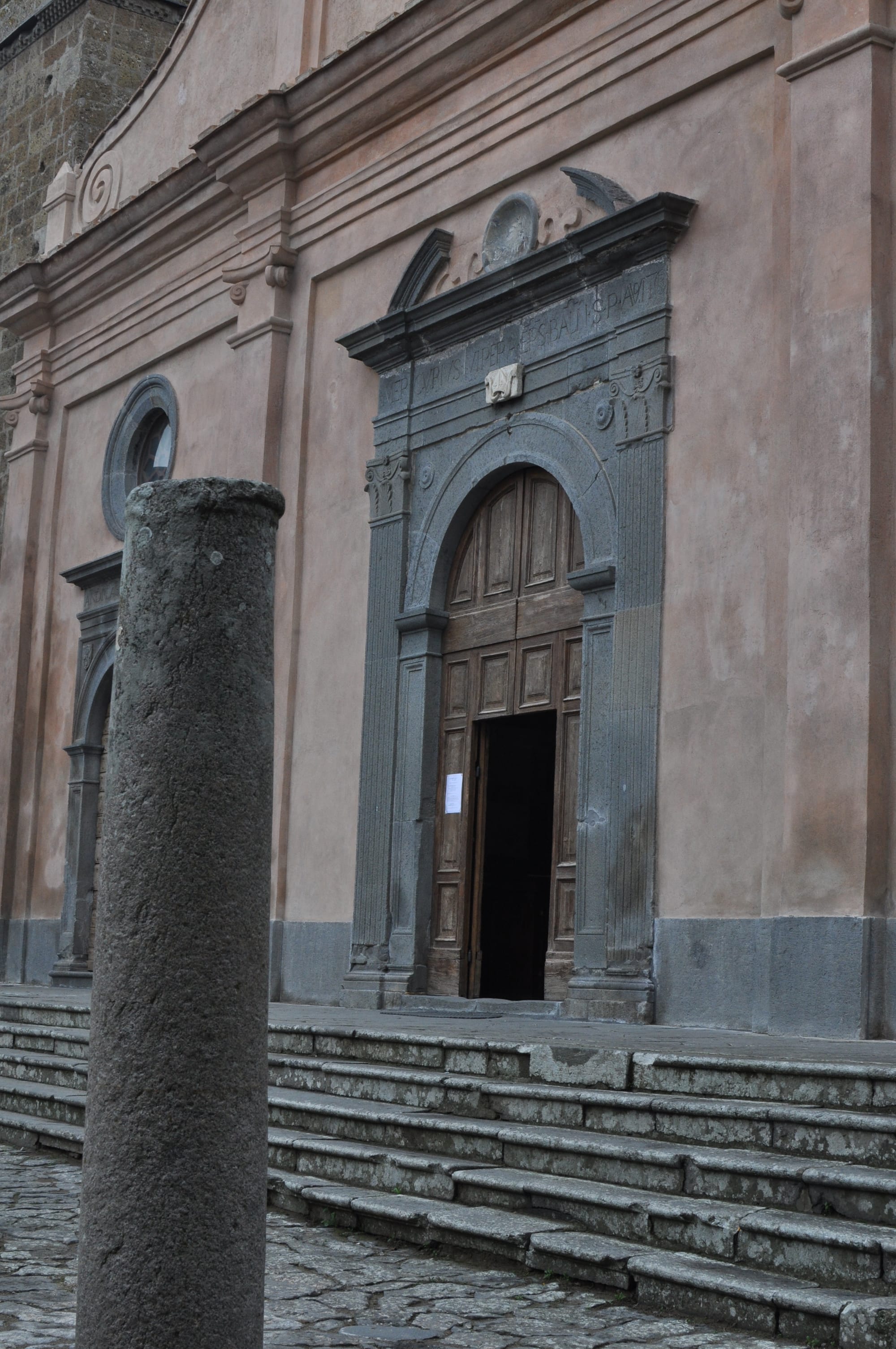 A close-up of a weathered stone Roman column standing in the foreground, with the stone steps and arched portal of San Donato Church blurred behind it.