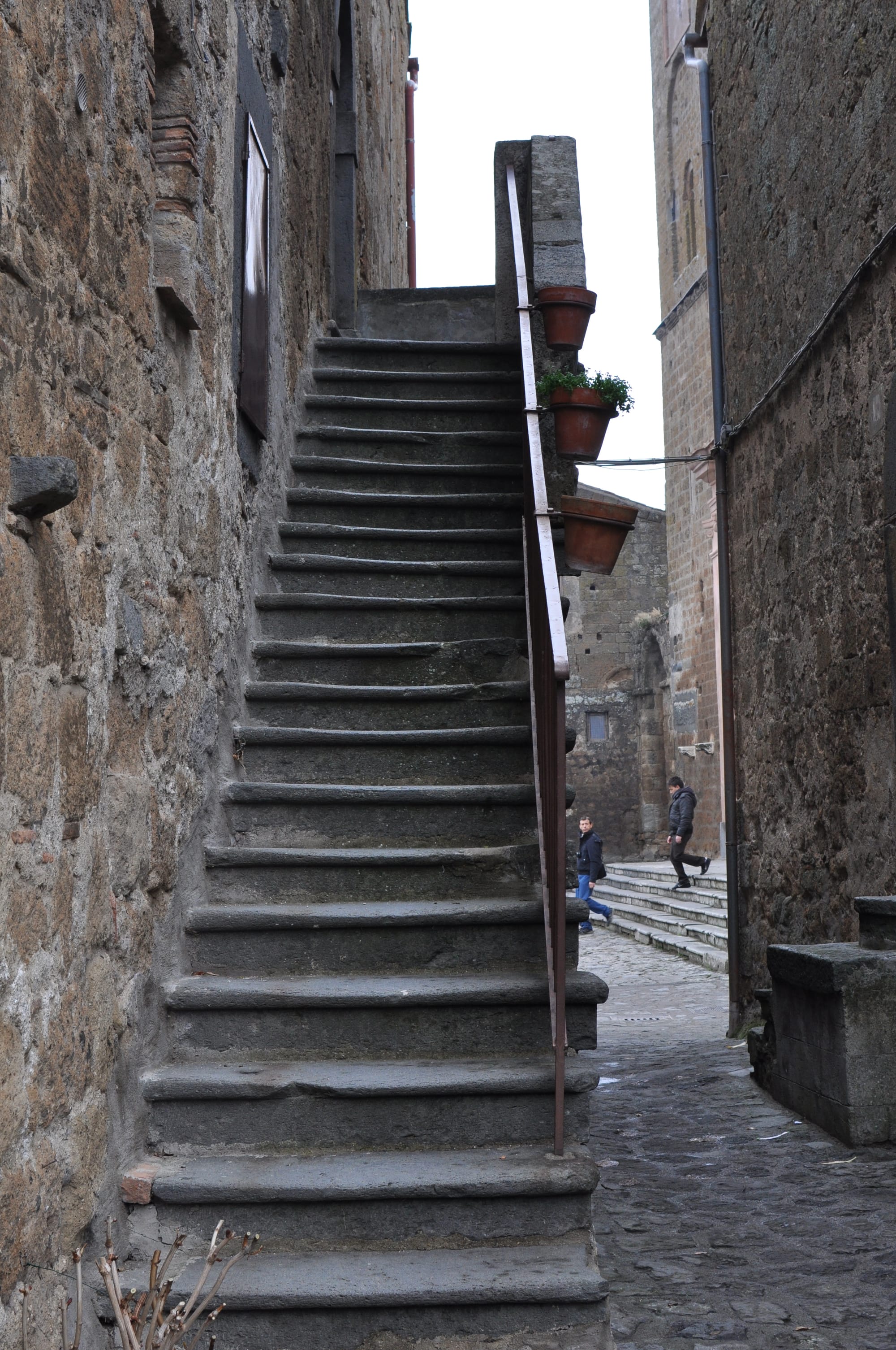 A steep, narrow stone staircase with a metal railing holding three terracotta pots climbs up along a rustic stone building wall in an alleyway.