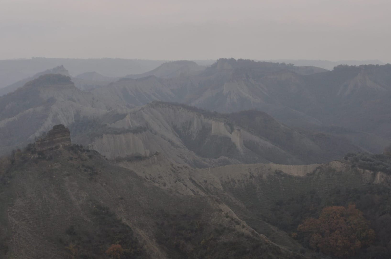 A hazy, wide view of rolling grey clay calanchi and eroded mountain ridges fading into a misty, overcast horizon.