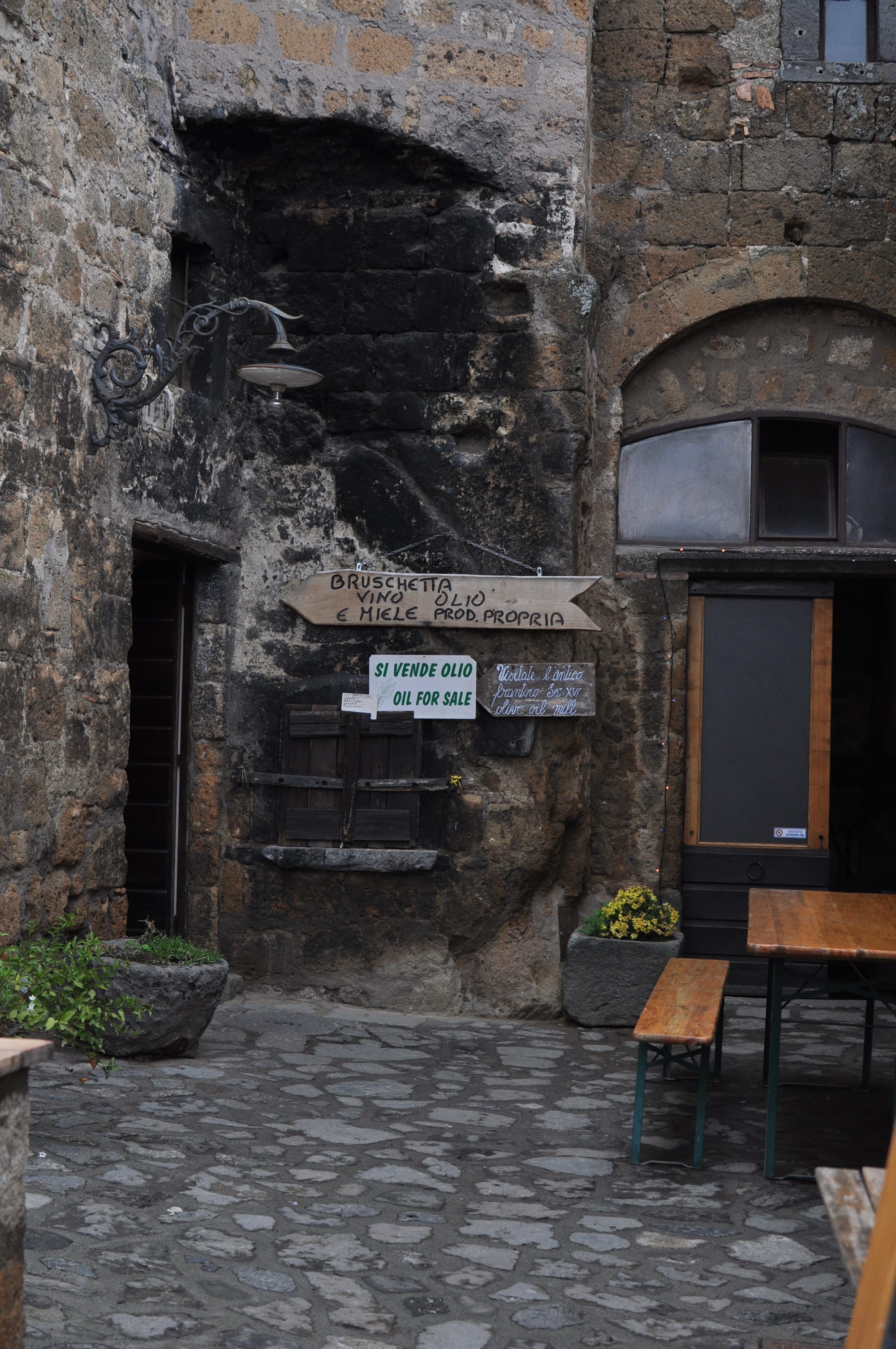 Wooden signs advertising local olive oil, wine, and bruschetta hang on an ancient, darkened stone courtyard wall next to a wooden bench.