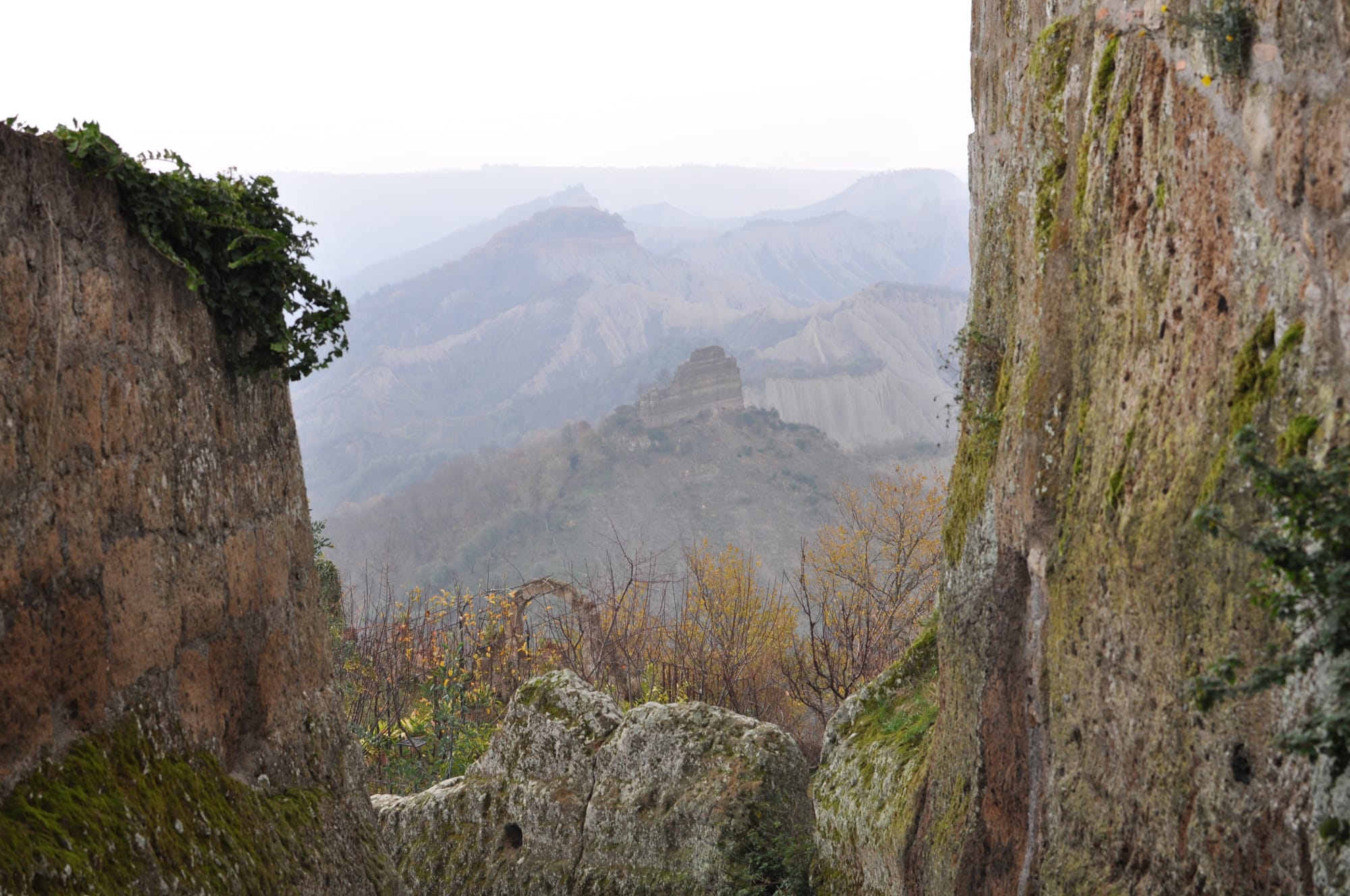 A view framed by two mossy stone walls looking out over a deep valley with a lone, ruined building standing on a distant ridge.