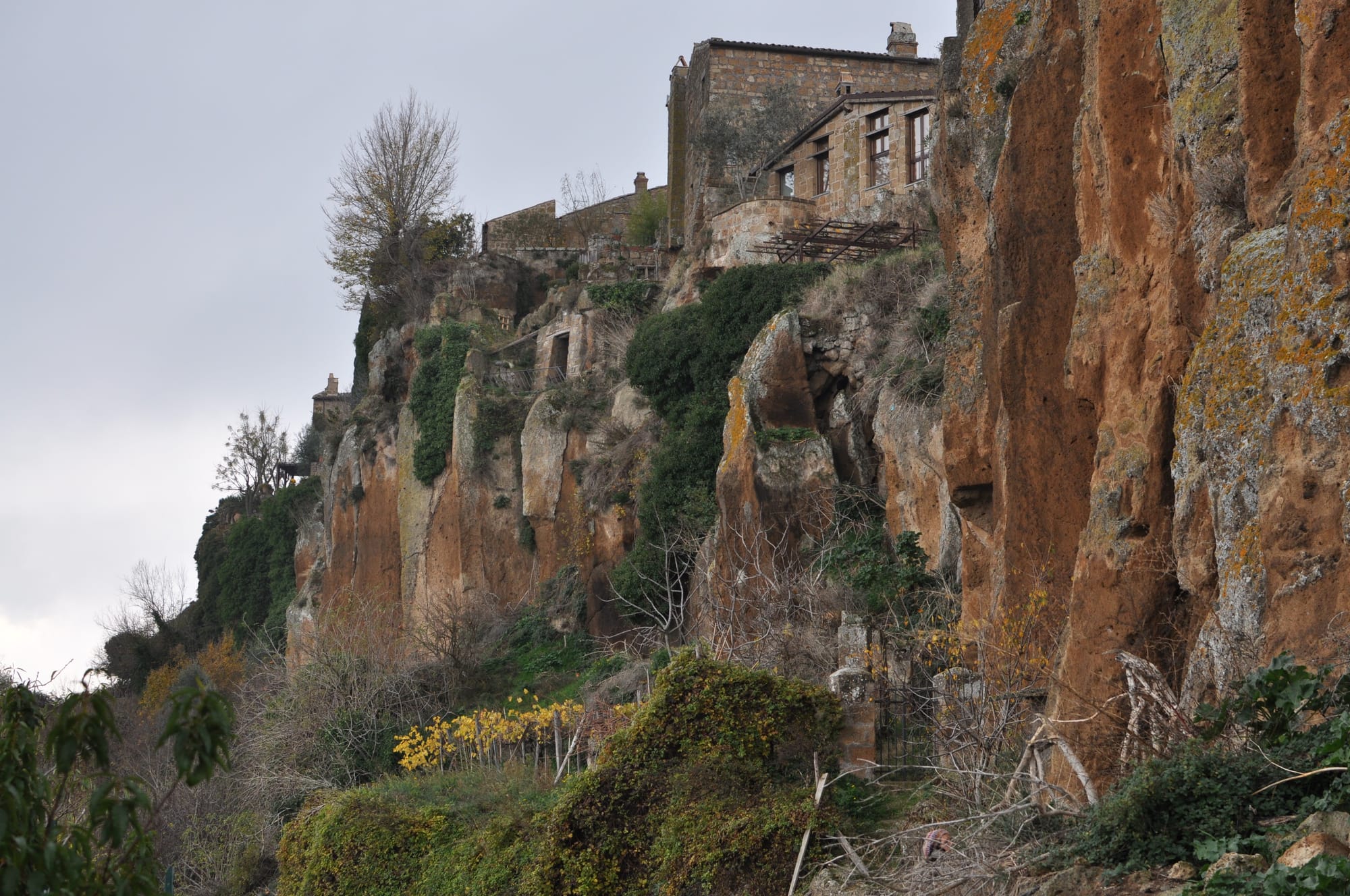Historic stone houses built directly onto the rugged edge of a high, sheer volcanic rock cliff face under a cloudy sky.