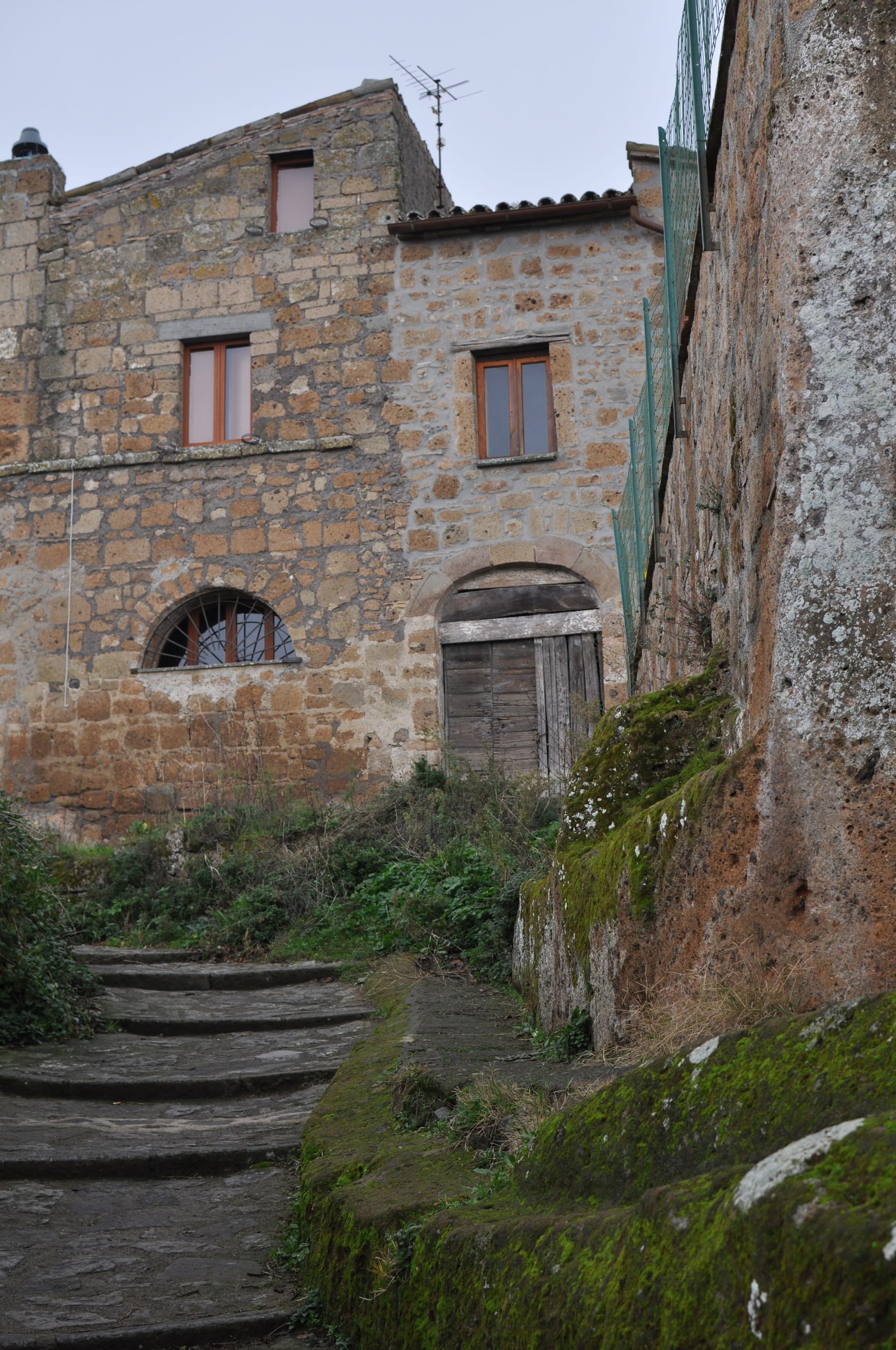 Mossy stone steps curve upward alongside a rugged rock wall leading toward traditional stone houses on an overcast day.