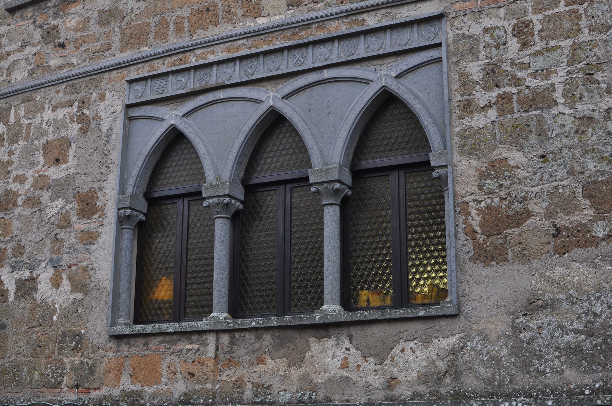Close-up of a decorative grey stone trifora window with three pointed arches and small carved heraldic shields on a rough tufa wall.
