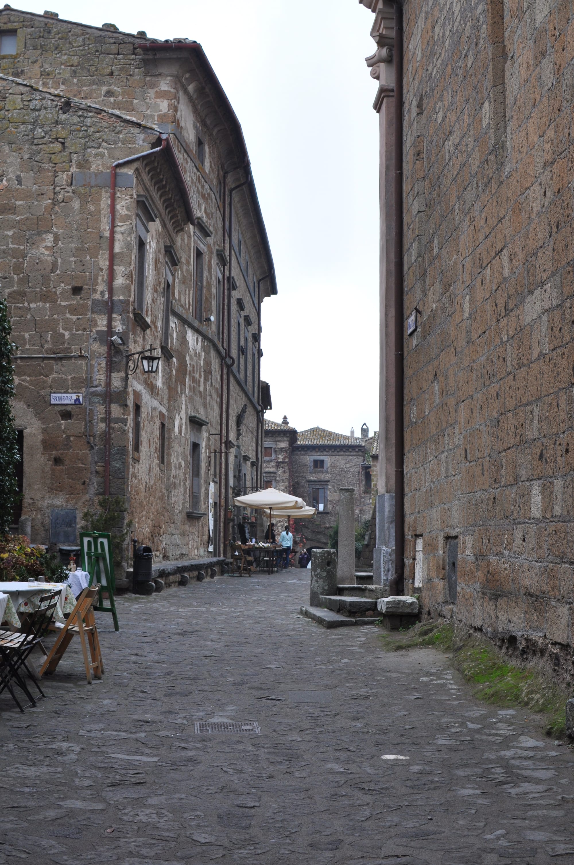 A wide view looking down a quiet stone street lined with historic buildings, featuring outdoor restaurant tables and umbrellas under a grey sky.