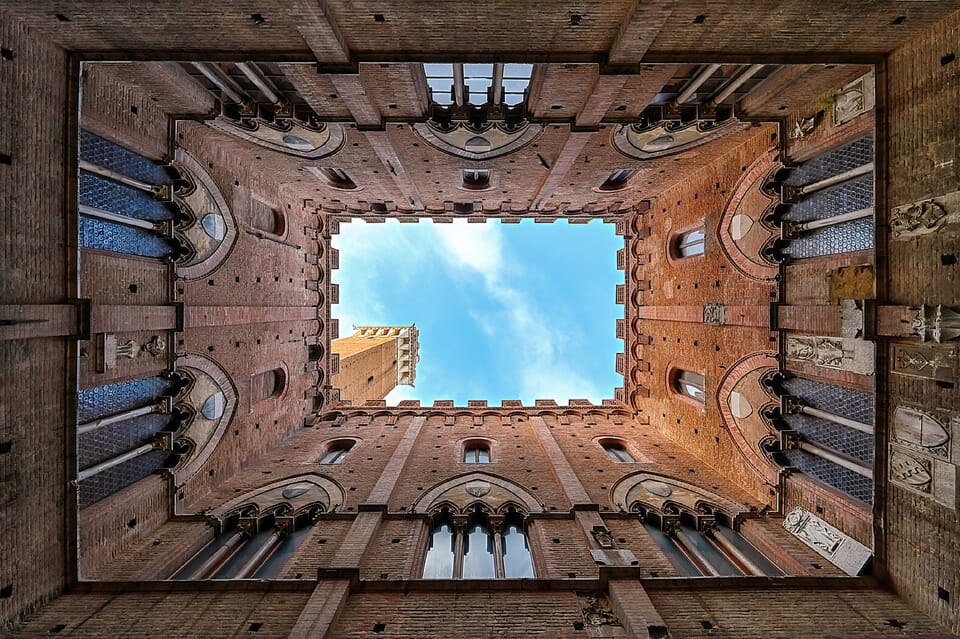A balanced, low-angle geometric shot from inside the square brick courtyard of the Palazzo Pubblico, looking straight up as the Torre del Mangia rises symmetrically into a clear blue sky.