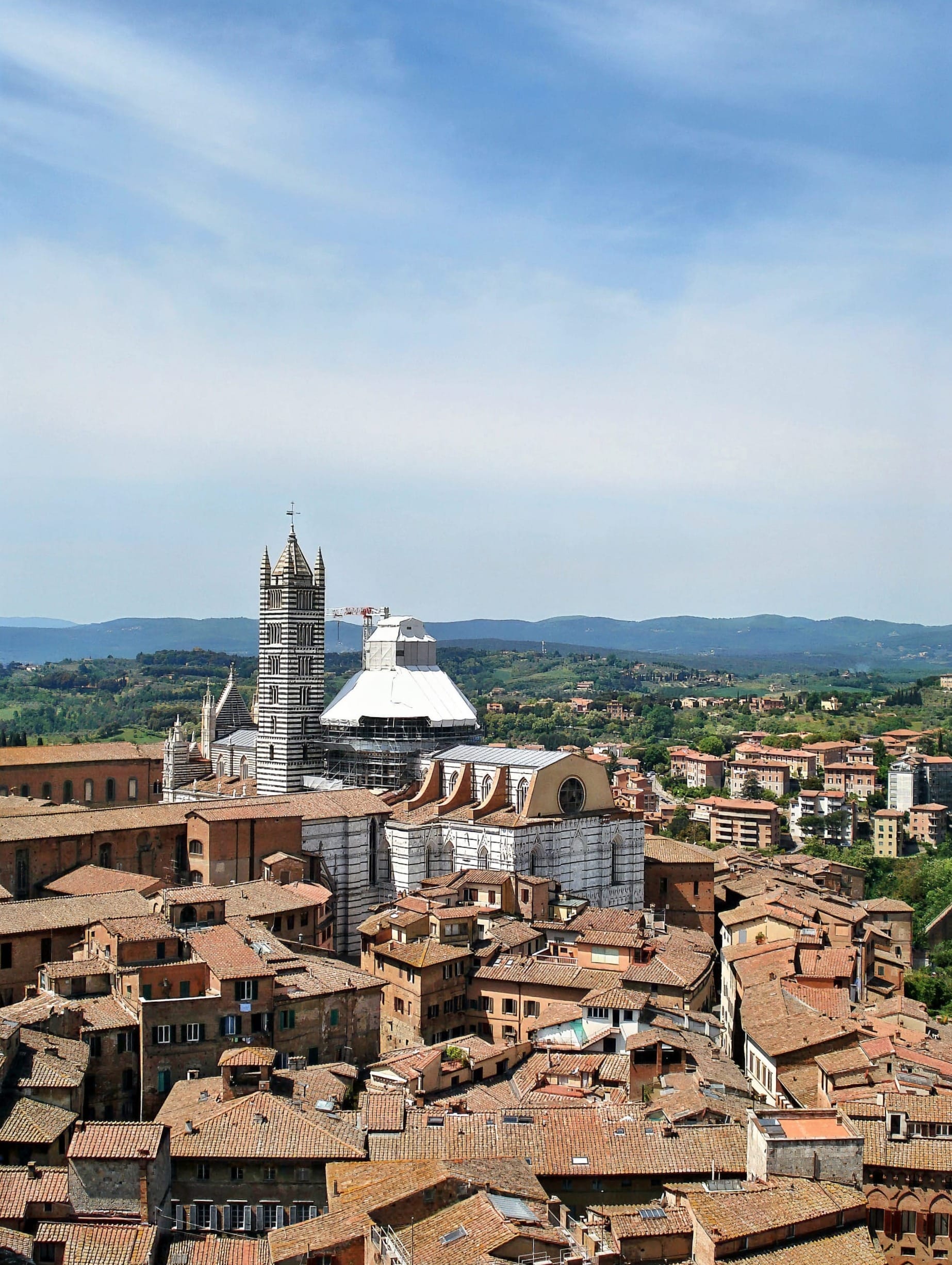 Panoramic view over the terracotta rooftops of Siena from the Torre del Mangia, with the Cathedral's striped facade and bell tower rising above the city.