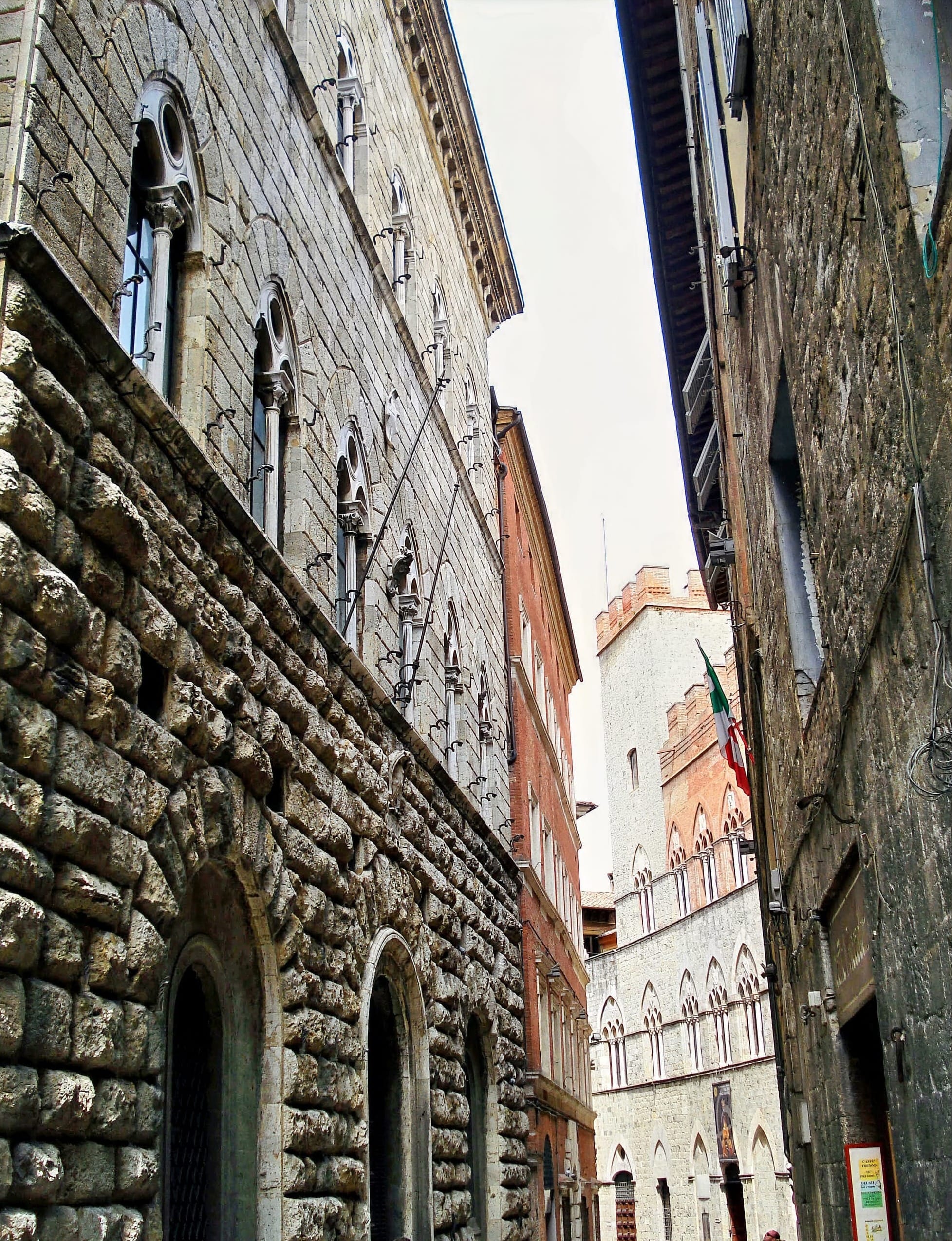 Close-up perspective of Via di Città in Siena showing the contrast between heavy rusticated stone blocks and red brick facades.
