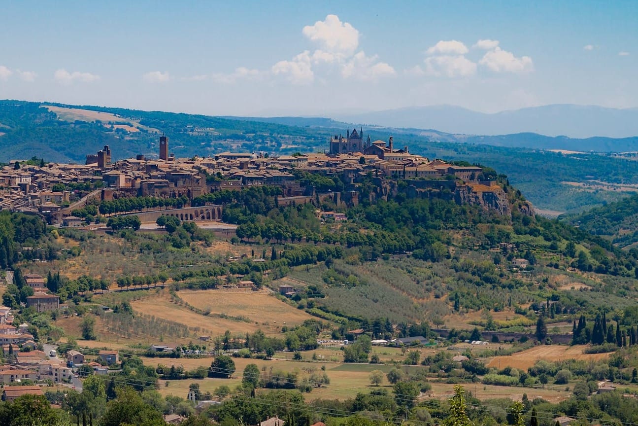 Wide panorama of the historic city of Orvieto perched on a sheer, vertical volcanic tufa cliff, isolated amidst the rolling green fields of the Umbrian countryside.