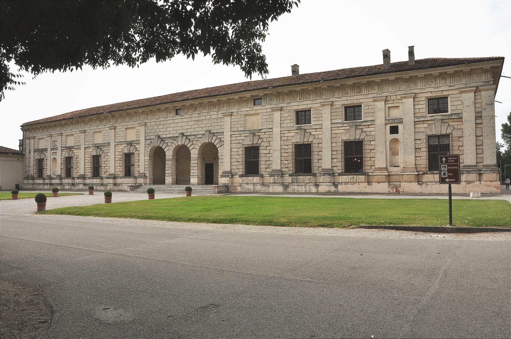 Symmetrical wide shot of the exterior facade of Palazzo Te in Mantua under an overcast sky, showing a rustic base and smooth pilasters.