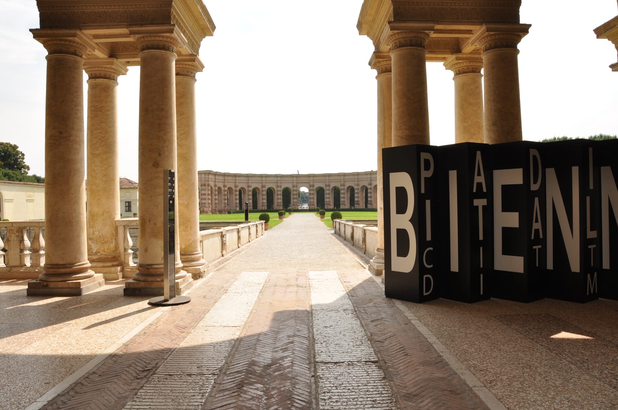 Interior perspective from a shaded classical loggia, looking past stone columns toward a massive, distant semicircular arcade across a sunlit garden.