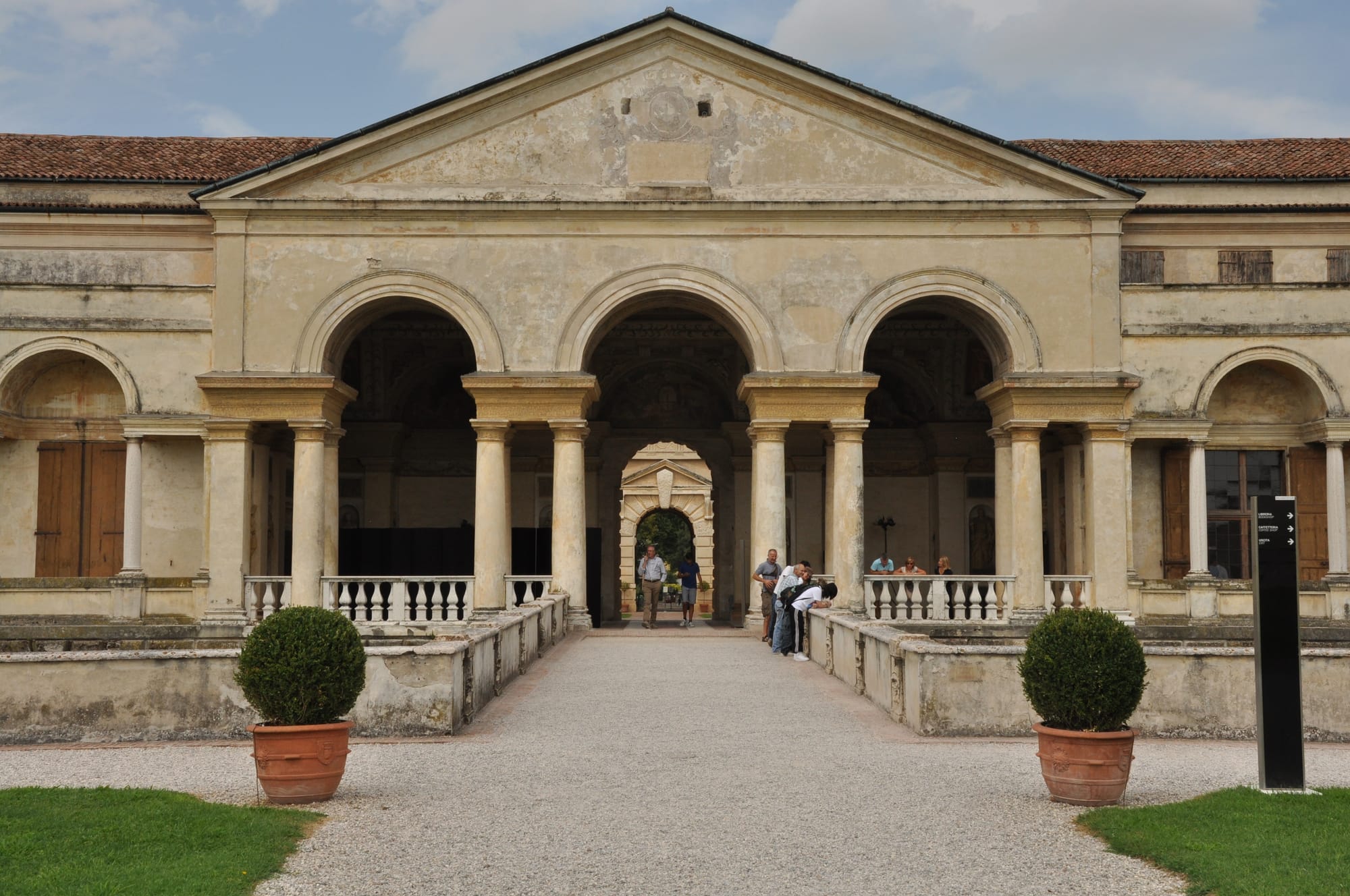 Front view of the grand triple-arched loggia at Palazzo Te, featuring paired classical columns, a weathered triangular pediment, and a gravel path flanked by potted boxwoods.