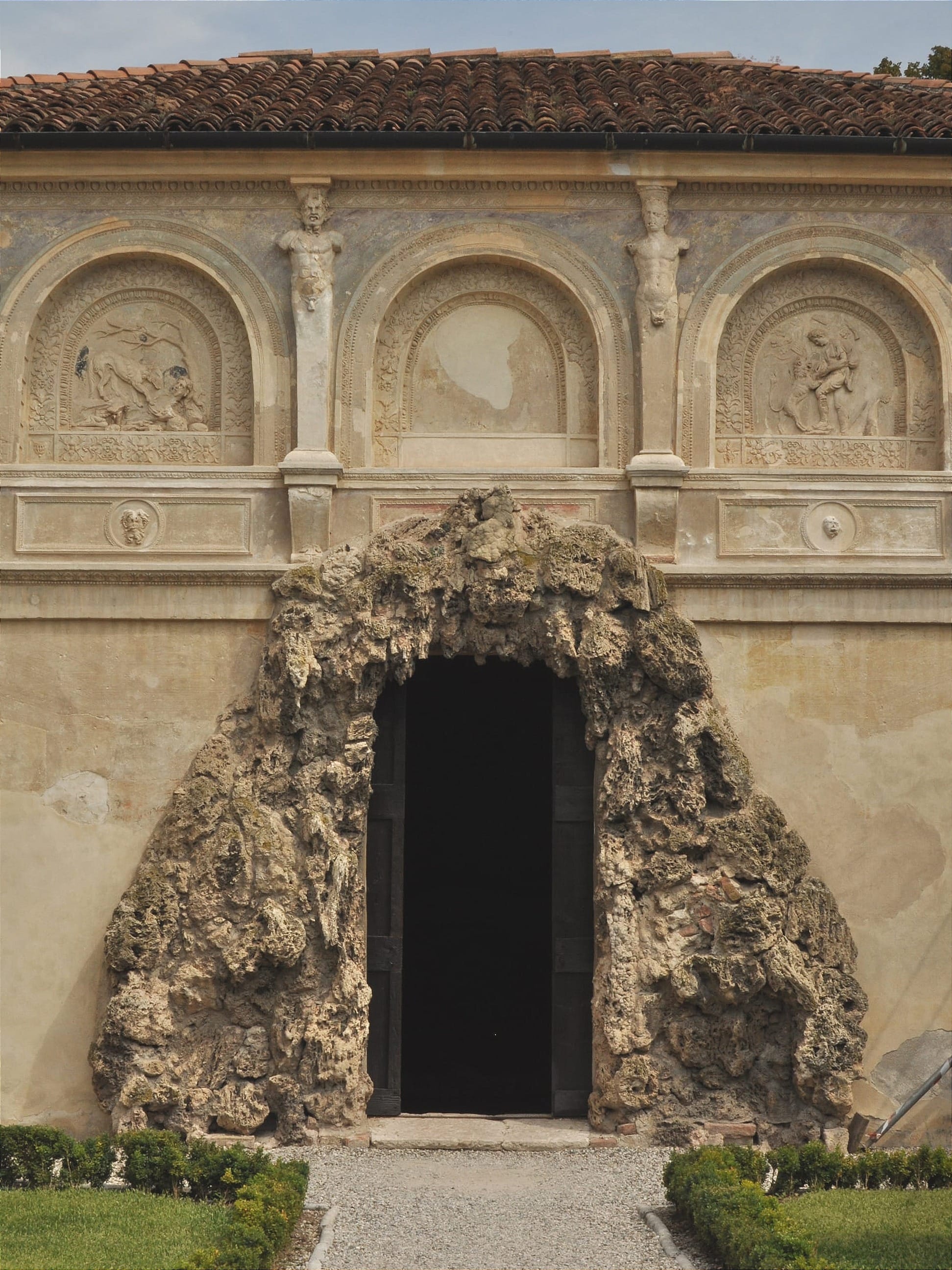 Symmetrical shot of a dark doorway framed by rough, porous rustic rockwork resembling a natural cave opening, set into a smooth plaster wall within a manicured garden.