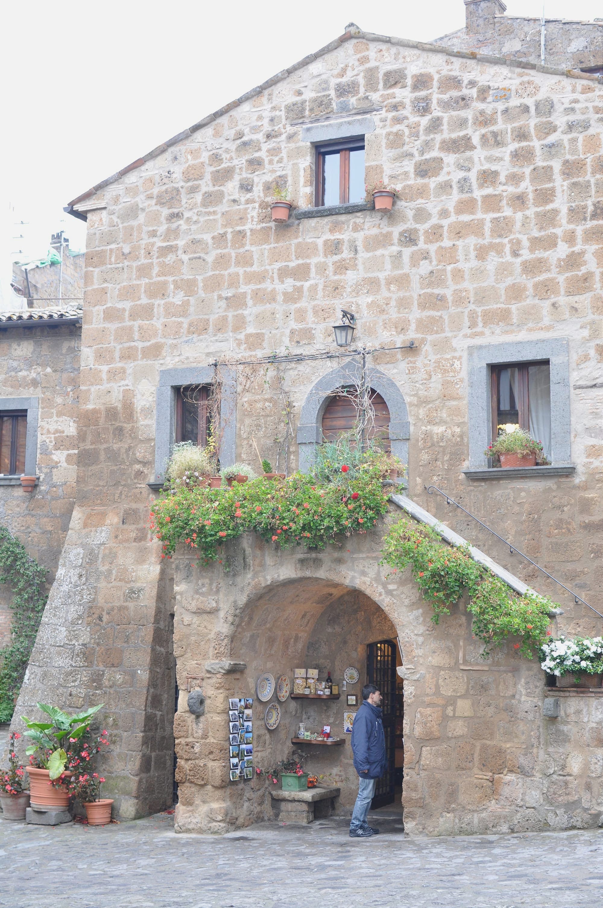 An ancient stone house with a large ground-floor arch framing a small shop entrance, decorated with climbing green plants and flower boxes on the windows.